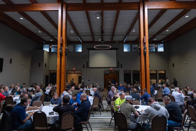 Large group of people seated at round tables in a spacious conference room with wooden beams and a blank projector screen.