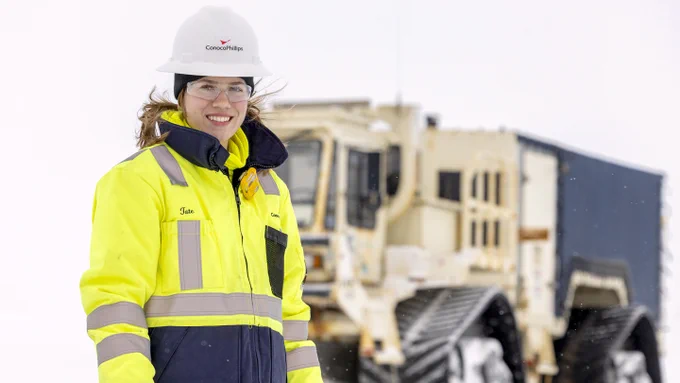 An individual is wearing a high-visibility jacket and a hard hat with the ConocoPhillips logo. They are standing in a snowy environment with a large, tracked vehicle in the background.
