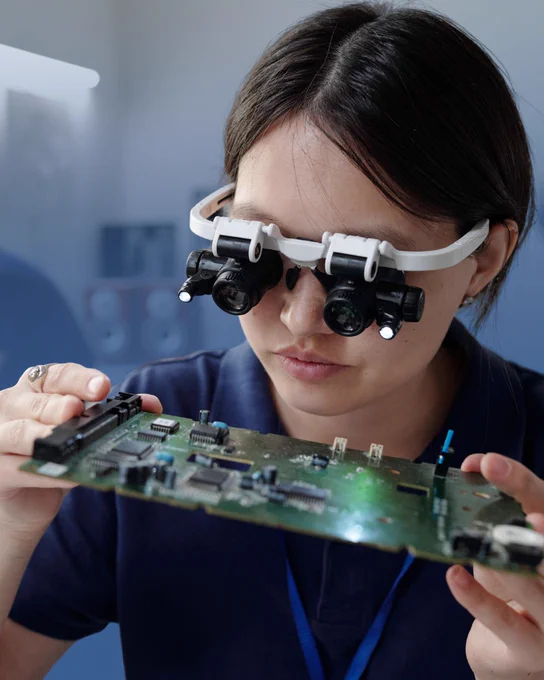 Person wearing magnifying glasses closely inspecting a circuit board in a lab setting.
