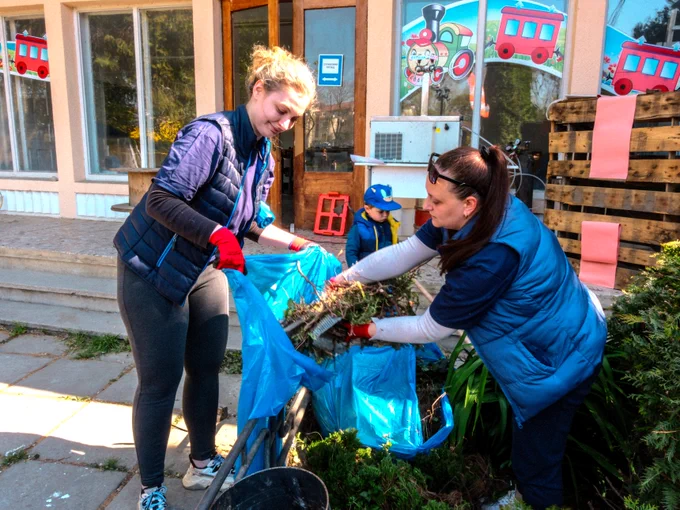 Two Uzhhorod employees wearing blue vests are cleaning outside a building at the Uzhhorod Children’s Railway. They are placing vegetation into blue bags. The building has a decorative window with cartoon train images.