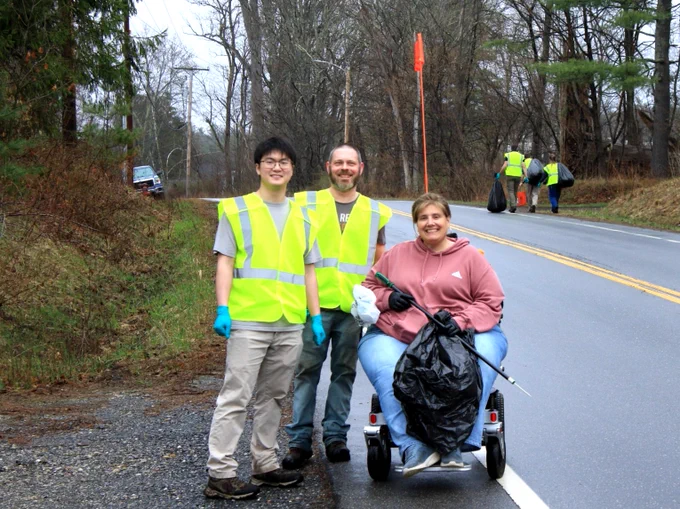 A group of three people in New Hampshire, wearing high-visibility vests, are volunteering to clean up the side of a road. One person is holding a trash bag while seated on a mobility scooter, and the others are standing, also holding cleanup tools. The road is flanked by trees, and other volunteers can be seen in the background.