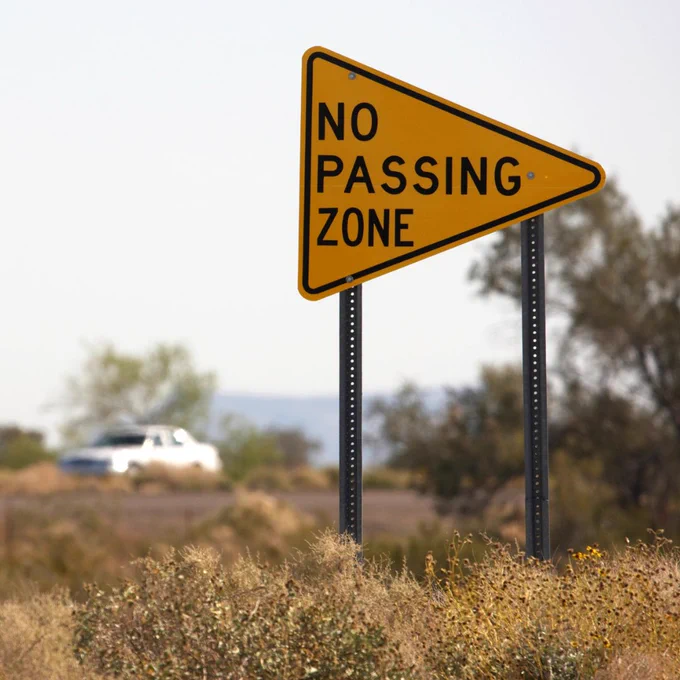 Image of a yellow road sign on the side of a rural road with car in background. Text on sign reads "No Passing Zone" 