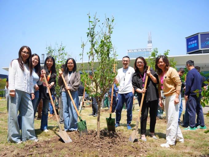 A group of people in Shanghai stand around a freshly planted tree, each holding a shovel. The scene is outdoors with a clear sky. 