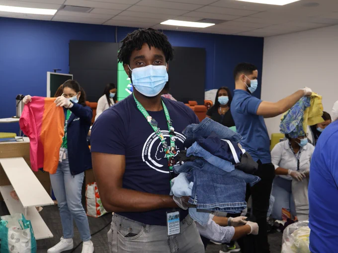 Employee poses in the Dominican Republic with a stack of pants as others in the background are folding and sorting clothes from a clothing drive.