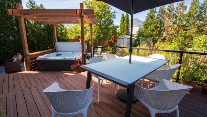 Backyard deck featuring a pergola-covered hot tub, outdoor dining table with chairs and umbrella, and surrounding greenery in a landscaped yard.