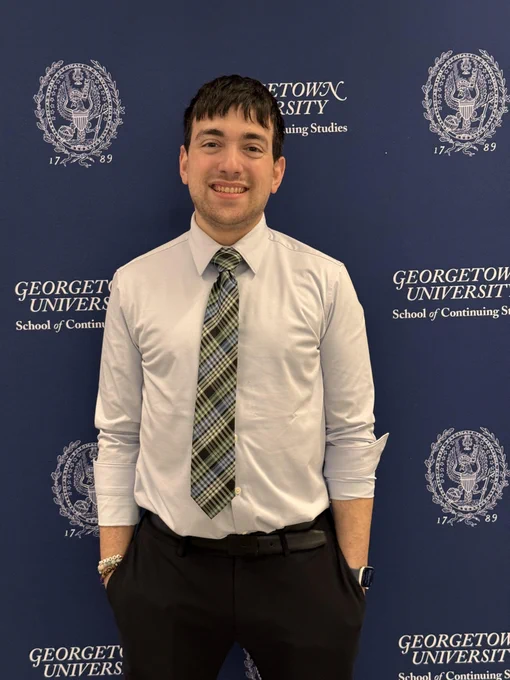 Farrell Moskow poses in front of a dark blue photo backdrop with the Georgetown University and Georgetown SCS logos.