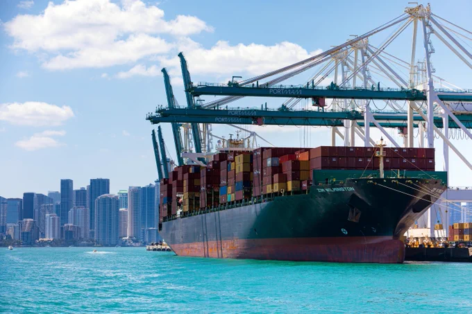A container ship named \"ZIM WILMINGTON\"@1 docked at Port Miami, loaded with containers, with cranes overhead against a backdrop of blue sky and city buildings. A small boat moves on the water.