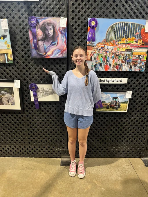 A young girl with brown hair, blue shorts and a blue shirt standing in front of a wall of artwork. She is pointing to a painting of a woman. The painting has a purple, Grand Champion, ribbon on it. 