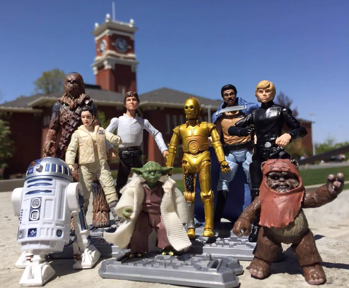 A lineup of small Star Wars action figures arranged on a gray plastic base sits outdoors in bright daylight. In the foreground and center stands a short, green, robed figure holding a cane, flanked by several humanoid and creature figures. To the left are a white-and-blue cylindrical droid in front and a tall, brown, fur-covered figure with bandolier straps behind a smaller figure in a light-colored outfit. Toward the middle are figures in a white vest, a shiny gold humanoid droid with circular chest detailing, and a figure wearing a dark cape and light pants. On the right stands a figure dressed head-to-toe in black and a small brown creature with a hood and raised arm. All figures face forward as if posing for a group photo. Behind them, slightly out of focus, is a red brick building with white trim and a clock tower, set against a clear blue sky and green lawn, suggesting a campus or civic setting.