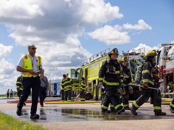 A group of firefighters in protective gear walks on a tarmac near several fire trucks, including a bright yellow one. One person in a reflective vest holds a clipboard. The sky is clear with a few clouds.