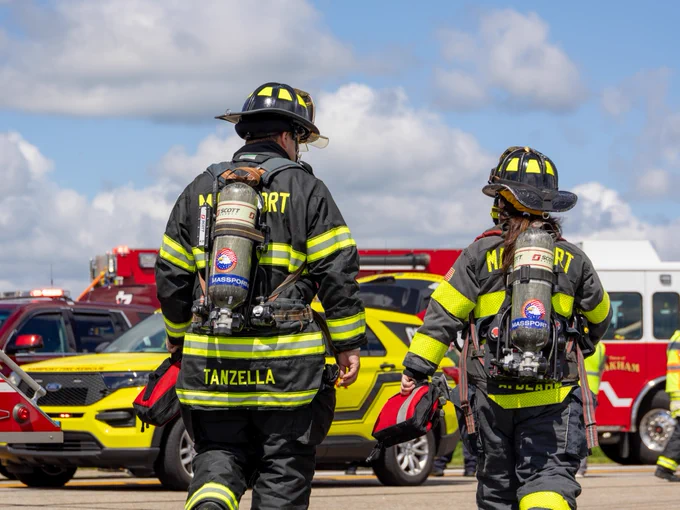 Two firefighters are walking away with their backs facing the camera. They are wearing full firefighting gear, including helmets and oxygen tanks. In the background, several fire trucks and emergency vehicles are visible. The sky is partly cloudy.