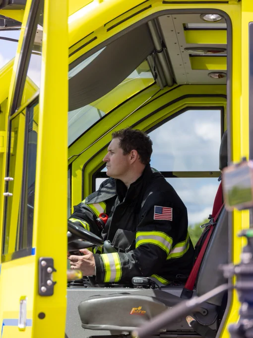 A person in a firefighting suit is seated inside a bright yellow emergency vehicle. The person appears focused, looking in the direction of the large front window.
