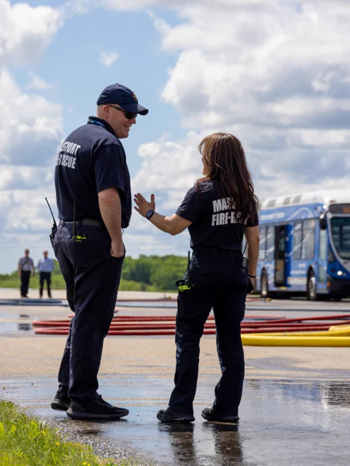 Two emergency responders are standing on a wet tarmac. They are engaged in conversation. A bus and coiled hoses are visible in the background under a partly cloudy sky.