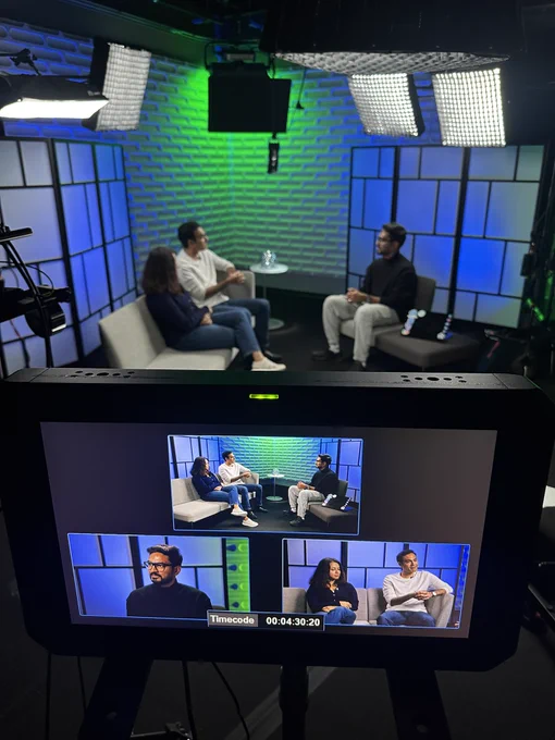 A studio setup with three individuals seated on couches having a discussion, with professional lighting and a green and blue background.