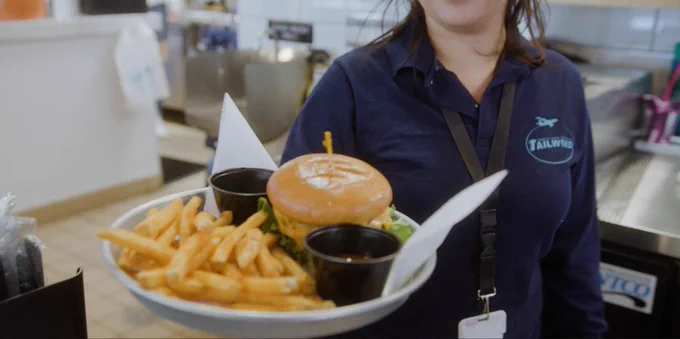 A Tailwinds employee serves a plate of a cheeseburger and fries over the lunch counter.