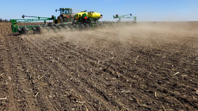 Large tractor pulling a planter machine seeding rows in a dry, dusty field under a clear blue sky.