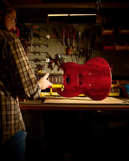 A person in the Gibson Custom Craftory is working on a red electric Gibson SG guitar body placed on a table. Various tools hang on a pegboard in the background. The lighting is focused on the guitar, highlighting its polished surface.