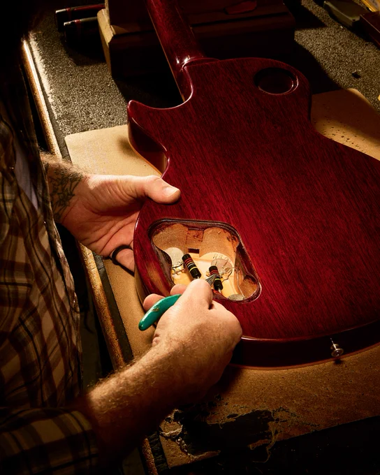 A person is working on a Gibson guitar in the Gibson Custom Craftory, focusing on the electronics inside the guitar's body. The guitar has a rich, dark wood finish.