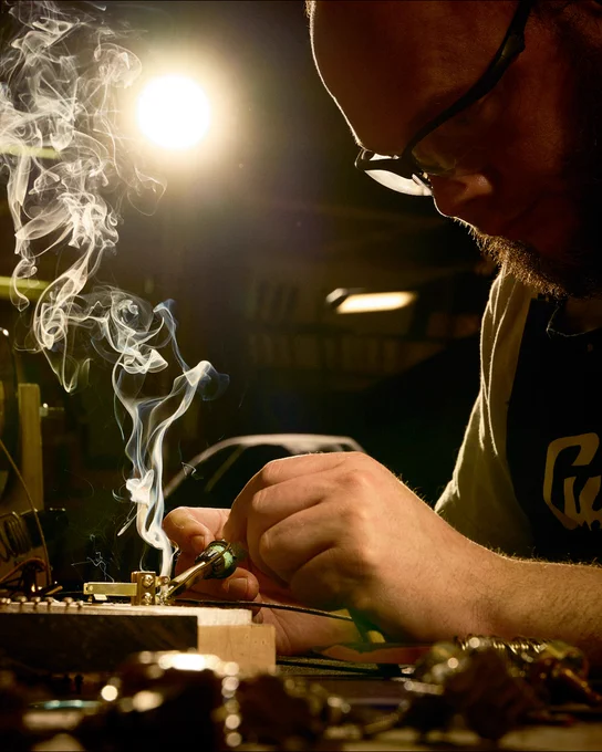 An individual is focused on soldering electronic components at a workbench in Gibson Custom Craftory, with smoke rising from the workstation. The scene is dimly lit, highlighting the concentrated task. A logo on the apron reads "Guild.