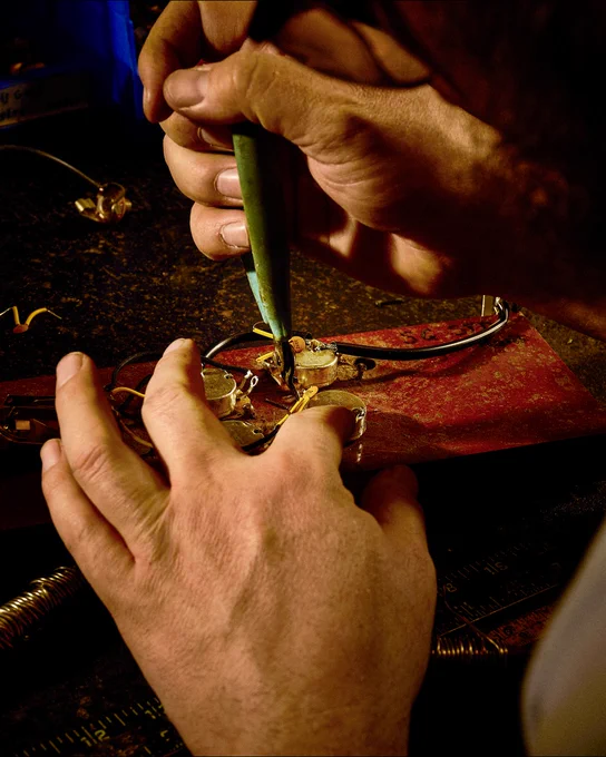 Close-up of hands carefully soldering electronic components on a circuit board in the Gibson Custom Craftory. A soldering iron is being used, with small tools and components visible.