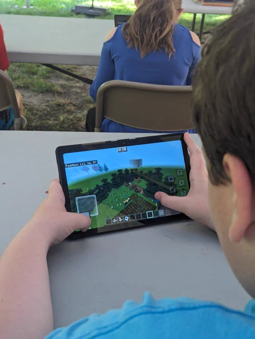 Back of a child using a tablet to build a homestead in Minecraft during an outdoor ranger program, with other participants seated at tables in the background.
