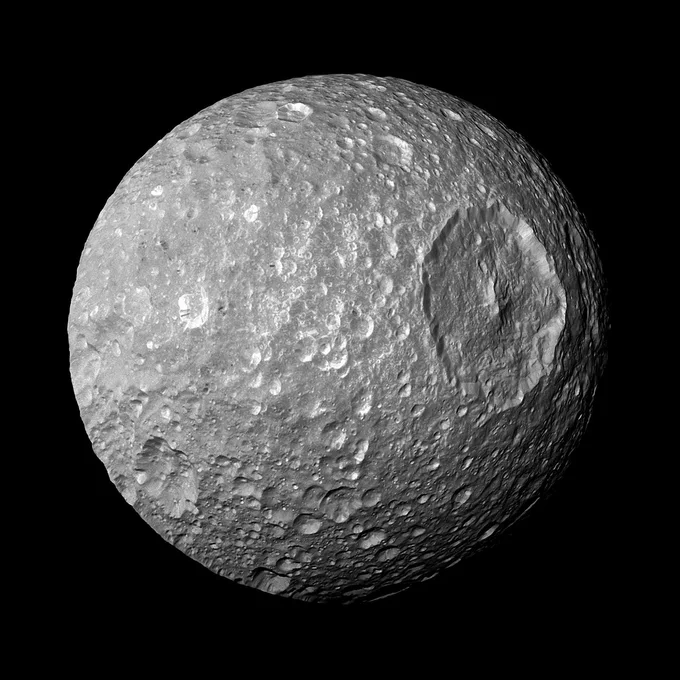 Saturn's moon Mimas appears in full phase against the darkness of space. Its heavily cratered surface is dominated by one large, circular crater that resembles the Concave Dish Composite Beam Superlaser aboard the Death Star in the Star Wars movies.