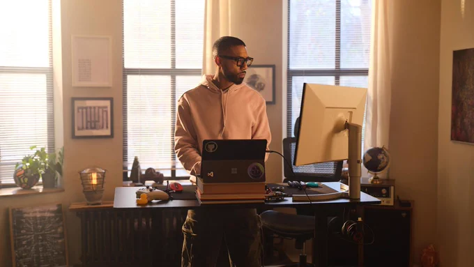 Photograph of a person standing at a desk with a laptop and a large monitor in a warmly lit room with large windows and beige curtains. The workspace includes various items such as a globe, plants, and framed pictures, suggesting a home office or creative work environment.​