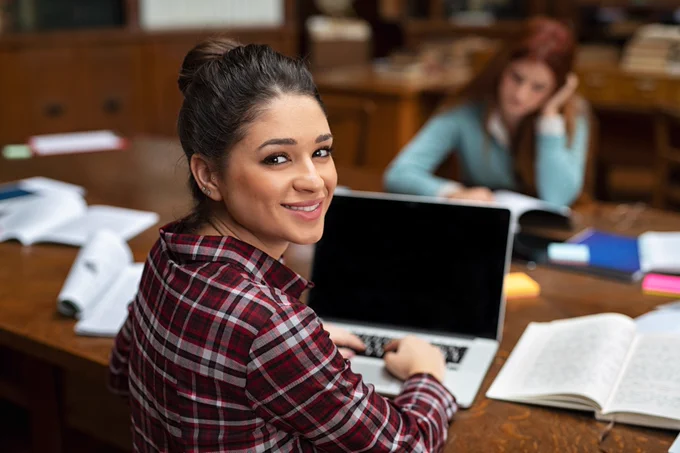 A high school student studying at her computer in a library turns around for a brief moment to smile at the camera. the table is wood. another student is in the background studying and out of focus.