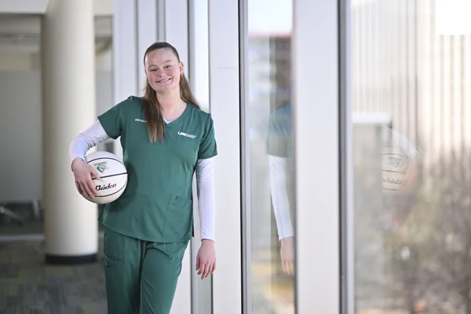 A person in green scrubs stands by a large window indoors, holding a white basketball with "Badin" written on it. The reflection of the person is visible in the glass.
