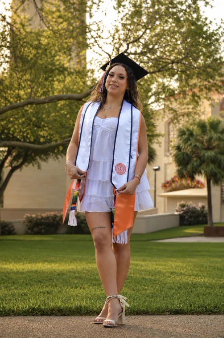 Carolina Wiltsey standing in a white dress with multiple stoles on and a graduation cap, smiling.