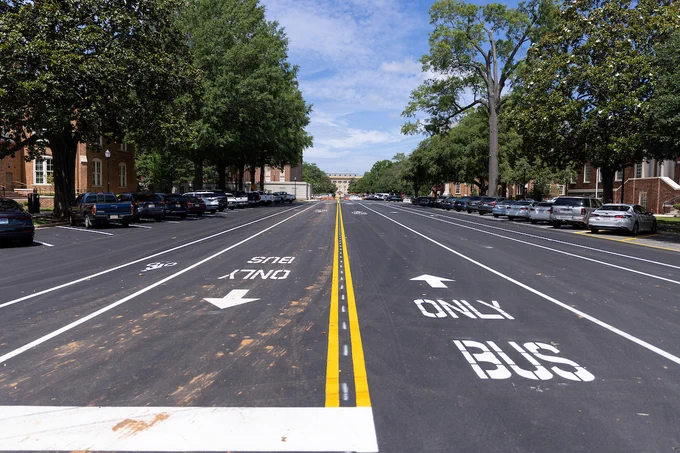 A wide street lined with parked cars and large trees on both sides. The road has clear markings, including arrows and the words "BUS ONLY" for lanes in both directions. The street leads to a distant building.
