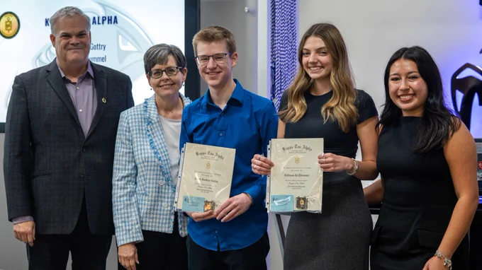 Five people stand together, with two holding certificates. One certificate mentions "Kappa Tau Alpha." They are in a room with a screen displaying a logo. All are dressed formally and appear to be at an awards event.
