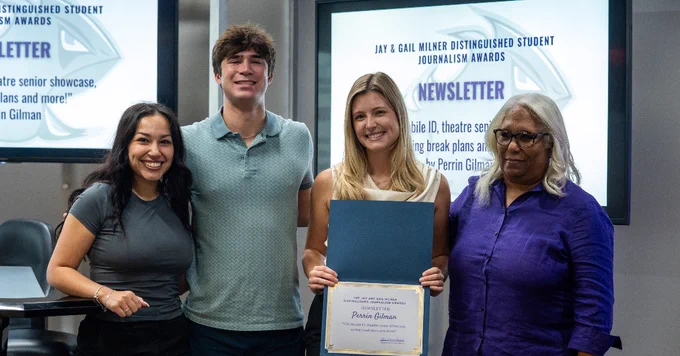 Four individuals are standing together indoors. One person is holding a certificate. In the background, a screen displays text about journalism awards and states "Newsletter".