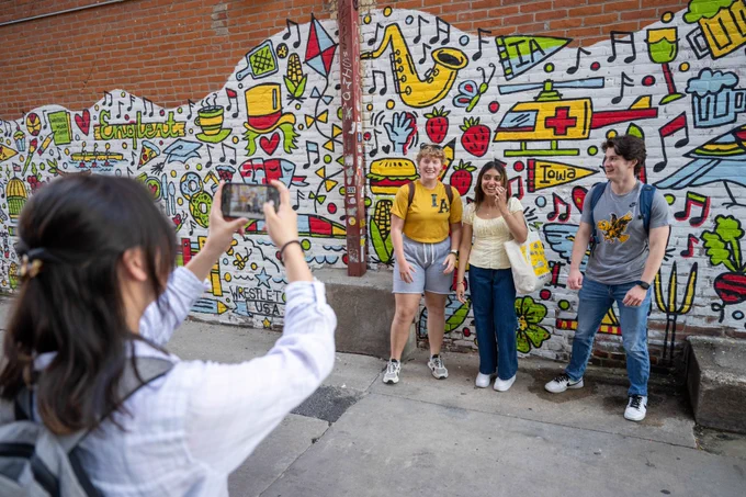 Students in front of a mural looking at a camera.
