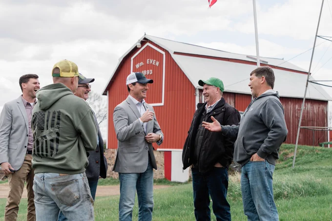 A group of people are standing and having a conversation outside near the Well-Even barn built in 1893. The barn is red with white trim and features are surrounded by a grassy area. Some individuals are wearing hats, and one person gestures while speaking.
