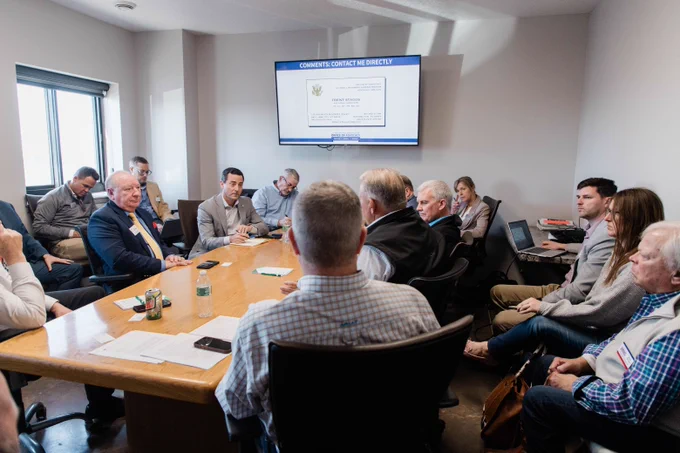 A group of people sitting around a conference table in a meeting room. A screen displays a slide titled "Comments Contact Me Directly" with further text underneath. Several individuals are looking towards the screen while others are engaged in conversation.