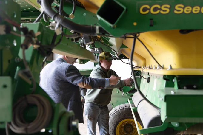 Two individuals are examining a large piece of green agricultural machinery labeled "CCS Seeder." They appear focused on adjusting or inspecting parts of the equipment. The setting is indoors with ample space around the machinery.
