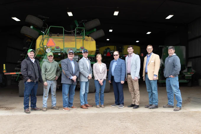 A group of people stand inside a spacious garage with farm equipment, including John Deere machinery, in the background. They're dressed casually and formally, with jackets and jeans. The group is positioned in the center of the well-lit space.