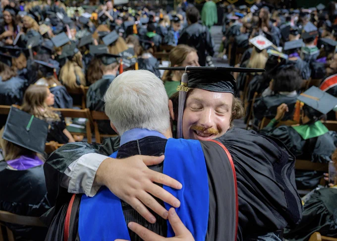 A graduate in their cap and gown embraces a person in academic regalia during a commencement ceremony. In the background, other graduates seated in rows are visible.