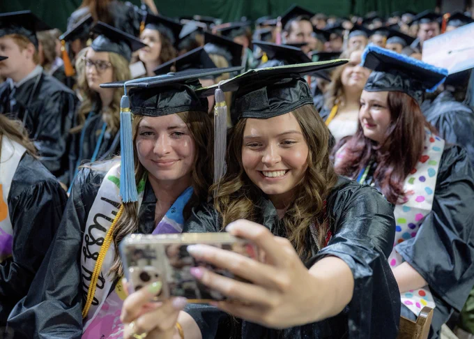 A graduate in their cap and gown takes a selfie with the person sitting to their left, who is also in their cap and gown. Rows of graduates can be seen behind them. 