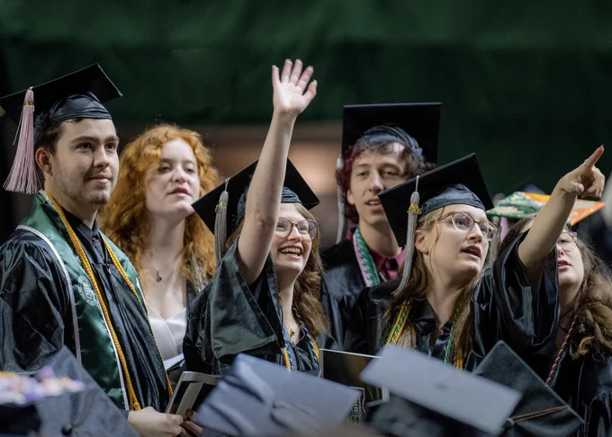 A graduate wearing their cap and gown stands and smiles while waving to someone in the audience. Another graduate to their right is pointing in the same direction. There are other graduates standing and the tops of the caps of those sitting in the foreground can be seen as well. 