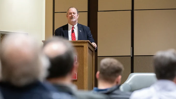 A man in a navy suit and red tie speaks at a wooden podium bearing a red University of Nebraska "N" logo. He faces a seated audience, visible in the blurred foreground. The setting is a lecture hall with neutral-toned walls.