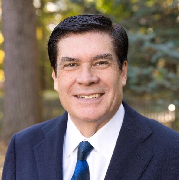 A professional headshot of Horacio Espinosa, a man with dark hair, wearing a dark suit, white dress shirt, and a blue patterned tie, smiling against a light blue background.