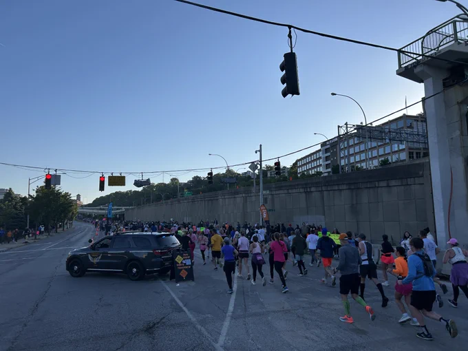 A marked OSHP Durango is in the middle of the road helping keep runners to one side of the road. Next to he Durango, is a sign that says, "Run today. Serve tomorrow." There is a QR code to join OSHP. Also there other words of encouragement for the runners. 