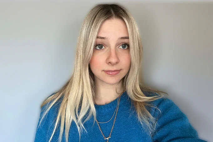 Carleton University journalism student Zoe Pierce takes a selfie. She has blonde hair and wears a blue sweater against a grey wall backdrop. 