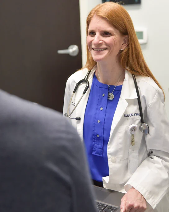 A doctor with long red hair wears a blue dress and white coat. She smiles and sits facing a patient in an exam room.