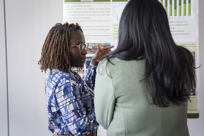 Two individuals stand in front of a presentation board displaying text and graphs. One gestures toward the board, possibly explaining the content. The setting suggests a professional or academic environment.