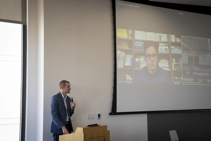 A person stands at a podium in a conference room, speaking into a microphone. On a large screen beside them, another individual is participating in a video call. The background of the screen shows shelves filled with books.