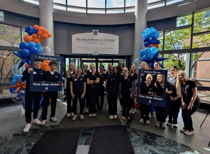 A group of people standing in a medical facility lobby holding blue and orange balloons, wearing black shirts with organization logo.
