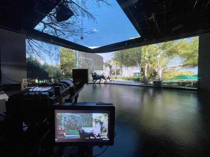 A high-tech studio with large screens display a scenic outdoor view featuring trees and greenery. A camera setup is visible in the foreground, capturing the scene displayed on the screens.
